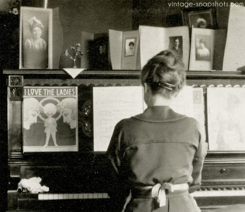 Vintage snapshot of woman playing piano, with sheet music and family portraits on piano